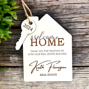 May include: White wooden house-shaped ornament with a key attached. The ornament says "Welcome Home" and "Thank you for trusting me with your real estate business. Keith Flanagan, Real Estate".
