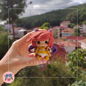 Op de afbeelding: Een gehaakte amigurumi-pop met rood haar, gele kleding en paarse vleugels. De pop wordt in een hand gehouden met een wazige achtergrond van huizen en bomen. De tekst "CHICKURIMI" is zichtbaar onder de pop.