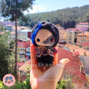 May include: A crocheted amigurumi doll with black hair, a blue and grey face, and a black and purple outfit. The doll is being held in a hand with the text "CHICKURIMI" visible below the doll.