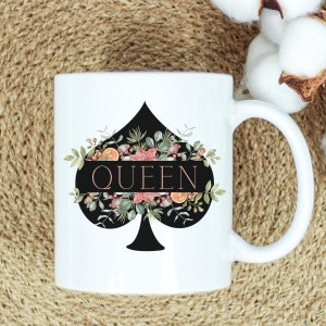 May include: White ceramic mug with a black spade design and floral accents. The word "QUEEN" is written in rose gold lettering.