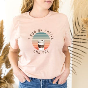 May include: A pink t-shirt with a retro-style graphic that says "I run on coffee and BBC". The graphic features a coffee cup with a brown lid and a white cup with a brown band. The graphic is set against a sunset-colored background with blue and green stripes.
