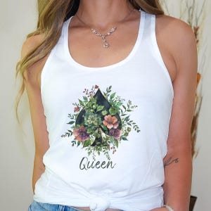 Może przedstawiać: Biały top typu racerback z centralną grafiką czarnego pika z talii kart, wypełnionego różnymi zielonymi liśćmi i kolorowymi kwiatami. Słowo Queen jest nadrukowane czarną kursywą bezpośrednio pod grafiką pika.