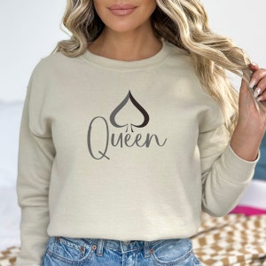 May include: A beige sweatshirt with a black and gray Queen of Spades graphic. The word "Queen" is written in a cursive font below the spade symbol. The sweatshirt has a crew neck and long sleeves.