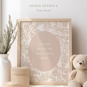 以下が含まれることがあります： ピンクの花柄のデザインと中央に「CUSTOM QUOTE OR BIBLE VERSE HERE」というテキストが書かれた円形が描かれた額装されたプリント。プリントは、白い花瓶、木製の箱、テディベアのあるテーブルの上に置かれています。