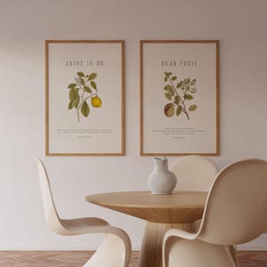 Op de afbeelding: Twee ingelijste prints met botanische illustraties en bijbelteksten. De prints hangen aan een witte muur boven een houten tafel met twee witte stoelen. De linker print zegt "Abide in Me" en de rechter print zegt "Bear Fruit".