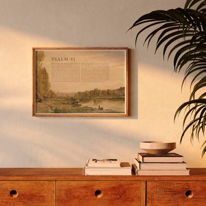 May include: A framed print of Psalm 91 with a landscape scene of a river and trees. The print is hanging on a beige wall above a wooden dresser with books and a bowl on top.