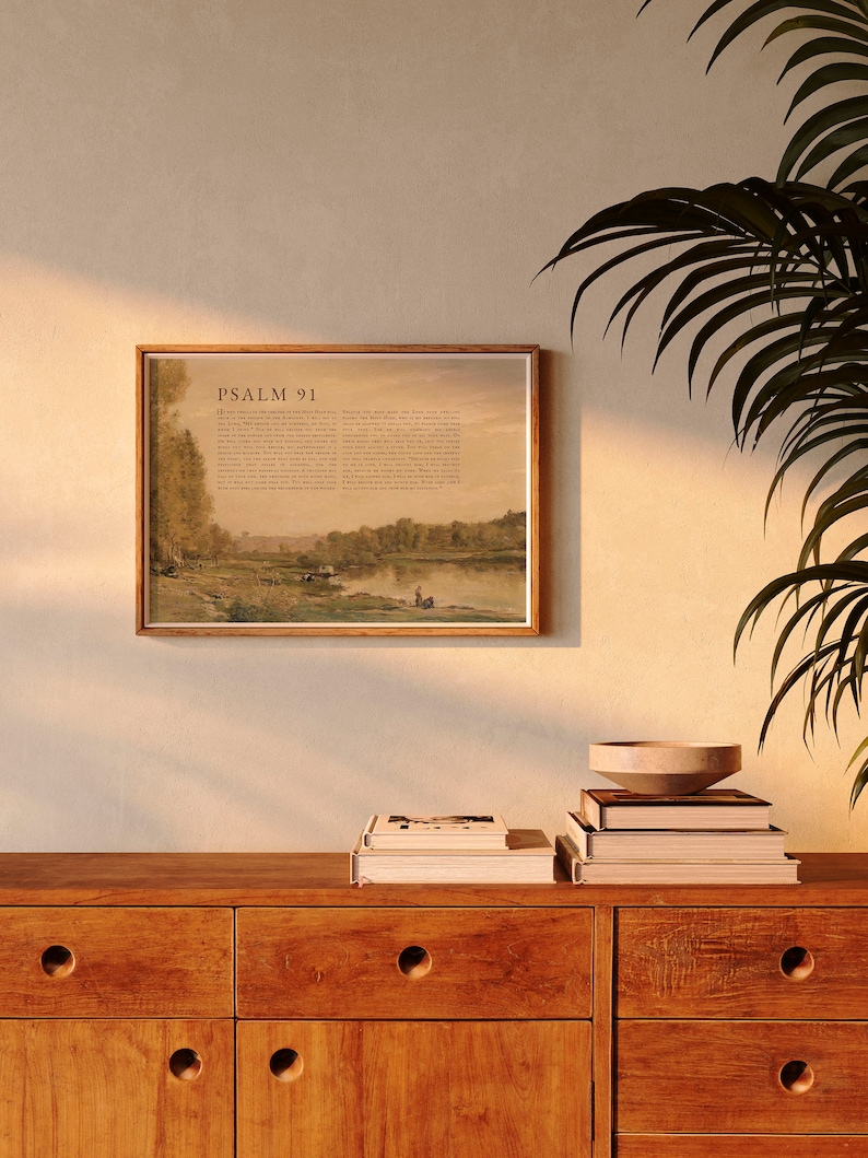 May include: A framed print of Psalm 91 with a landscape scene of a river and trees. The print is hanging on a beige wall above a wooden dresser with books and a bowl on top.