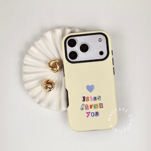 May include: A cream-colored phone case with the text "Jesus loves you" in colorful letters. A blue gingham heart is above the text. The case is next to a white, scalloped dish with two gold-colored knot earrings.