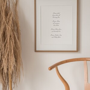 Może przedstawiać: Wydruk w ramie z jasnego drewna i białym tłem. Wydruk zawiera cytat religijny w ozdobnym piśmie: "Chwalcie Boga, od kt&oacute;rego pochodzą wszystkie błogosławieństwa. Chwalcie Go, wszystkie stworzenia na ziemi. Chwalcie Go w g&oacute;rze, wy, zastępy niebieskie. Chwalcie Ojca, Syna i Ducha Świętego."