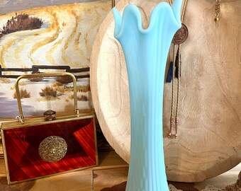 Pode incluir: Um vaso alto de vidro azul claro com uma borda ondulada. O vaso tem detalhes verticais canelados e uma base texturizada. Uma bolsa vermelha e dourada e uma pintura de paisagem estão no fundo.