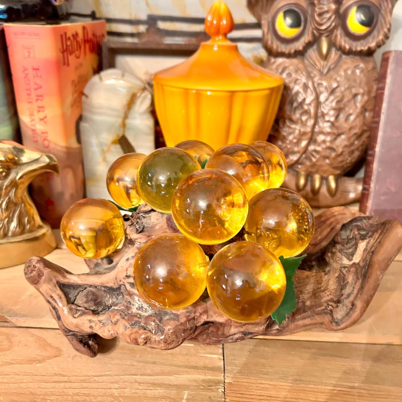 May include: A cluster of amber glass grapes on a piece of driftwood. The grapes are arranged in a bunch and have a realistic appearance.