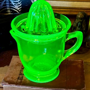 Peut inclure: Presse-agrumes en verre vert vif, composé d'une base en forme de pichet et d'un presse-agrumes. Il est doté d'une poignée et d'un bec verseur. Le verre présente un aspect texturé. Le presse-agrumes est posé sur un livre brun.