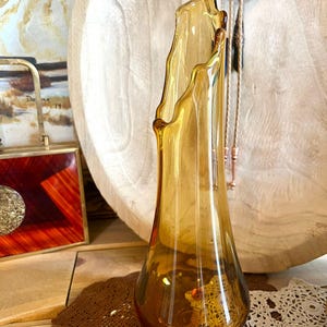 May include: Amber-coloured glass vase with an unusual, asymmetrical rim. The vase has a flared base and a slender, elongated body. The glass has a warm, golden hue, and the vase is likely vintage or antique. The vase is displayed on a wooden surface.