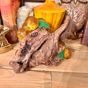 May include: A decorative piece featuring a cluster of amber glass grapes resting on a piece of driftwood with green leaves.