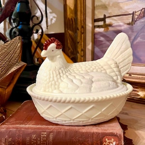 May include: Cream-colored ceramic hen-shaped covered dish. The hen has a detailed textured body, a red comb, and a white face. The dish sits on a decorative base with a diamond pattern. A vintage book is beneath the dish.