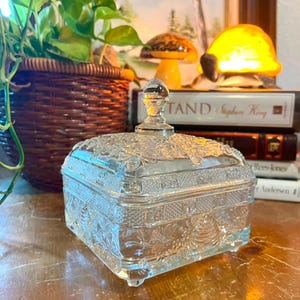 May include: A square clear glass trinket box with a removable lid featuring a decorative finial. The glass surface is embossed with intricate bee and honeycomb patterns, and the base is supported by four small feet at the corners.