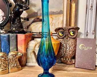 Pode incluir: Um vaso de vidro azul alto com um design de espiral. O vaso tem uma boca larga e uma base estreita. Está sentado em uma mesa de madeira com livros e outros itens decorativos.