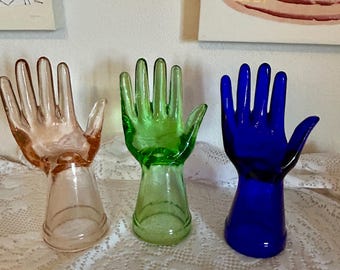 Peut inclure: Trois supports de bague en verre en forme de main. Les mains sont en rose, vert et bleu.