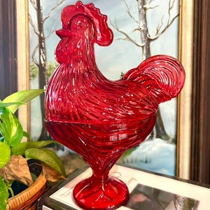 May include: A vibrant red glass rooster figurine featuring a detailed molded texture on the feathers and comb. The rooster stands on a circular pedestal base and is designed as a lidded dish with a horizontal seam separating the top and bottom halves.