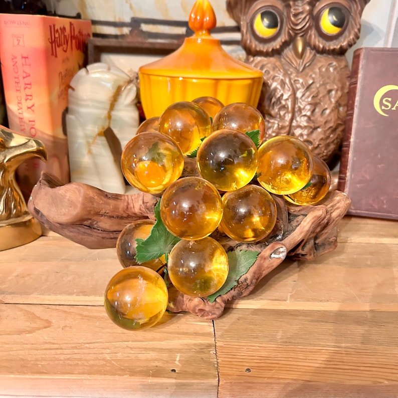 May include: A cluster of amber glass grapes on a piece of driftwood. The grapes are arranged in a bunch and have a realistic appearance.