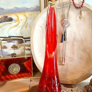 May include: A striking red glass vase, featuring a flared base and a narrow neck. The vase has a textured surface with small bubbles. It is displayed on a wooden surface, with other decorative items in the background. The vase's height is approximately 30 cm.