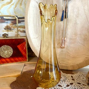 May include: Amber glass vase with a flared, scalloped rim. The vase has a slender, elongated body and a detailed base. The glass has a slight iridescence. A decorative piece for home decor.