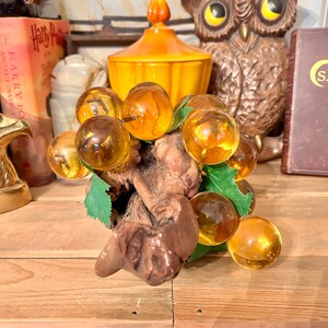 May include: A glass grape cluster with amber-coloured glass grapes on a wooden branch with green leaves.