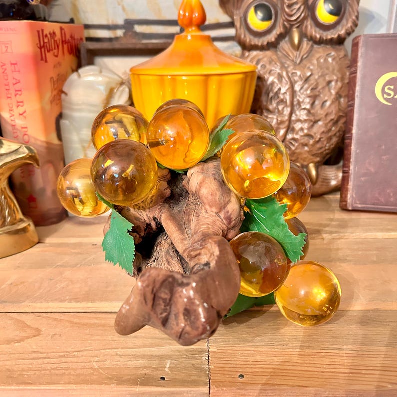 May include: A glass grape cluster with amber-coloured glass grapes on a wooden branch with green leaves.