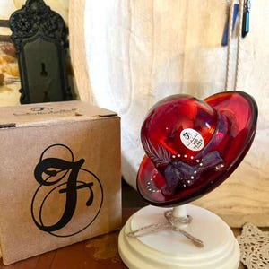 May include: A vibrant red glass hat-shaped decorative object on a white stand, tied with twine. A brown box with a large black letter "F" is to the left. The hat has a wide brim and a rounded top, with a label.