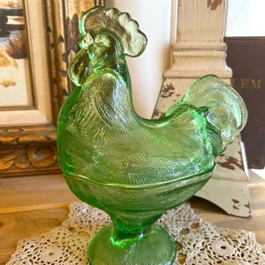 Puede incluir: Una figura de gallo de vidrio verde lima translúcido con un patrón de plumas moldeado detallado en el cuerpo, el cuello y la cola. El gallo se apoya sobre una base circular e incluye una tapa extraíble que forma la parte superior del ave.
