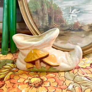 May include: A ceramic planter shaped like a shoe with a mushroom design. The planter is white with brown speckles and features three yellow mushrooms with green leaves.