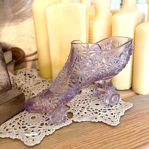May include: A lavender-colored glass slipper with a detailed starburst pattern, resting on small clear wheels. The decorative shoe sits on a cream-colored doily, with yellow candles in the background. A vintage-inspired decorative piece.