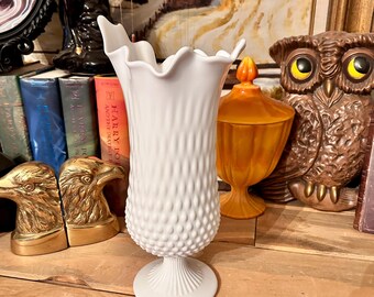 Puede incluir: Un jarrón de vidrio de leche blanco con una superficie texturizada y abollonada y un borde festoneado. El jarrón tiene un diseño clásico y elegante y es perfecto para exhibir flores u otros artículos decorativos.