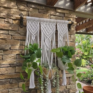 May include: A white macramé plant hanger with three sections, each holding a potted plant with green leaves. The macramé is mounted on a wooden dowel and hangs against a stone wall. A woven basket holds a trailing plant.
