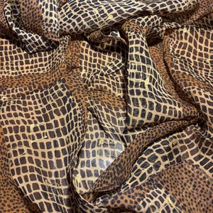 May include: A flowing fabric with a brown and black reptile-like pattern. The material has a textured appearance with a repeating square design. Hints of gold shimmer throughout the fabric, adding a touch of elegance.
