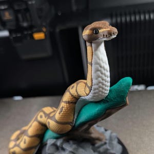 May include: A 3D printed figurine of a ball python with brown and tan patterned scales and a white underbelly. The snake is coiled around a green branch that is attached to a textured gray rock base, featuring a smooth black circular bottom.