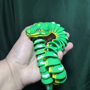 May include: A flexible, articulated toy snake featuring a vibrant green body with textured scales, bright yellow accents along the head and underside, and white markings. The segmented design allows for fluid movement and realistic posing.