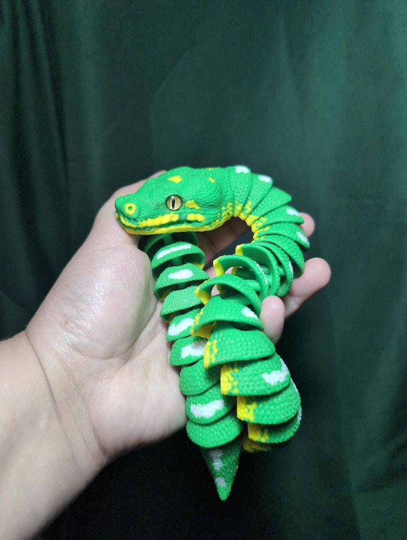 May include: A flexible, articulated toy snake featuring a vibrant green body with textured scales, bright yellow accents along the head and underside, and white markings. The segmented design allows for fluid movement and realistic posing.