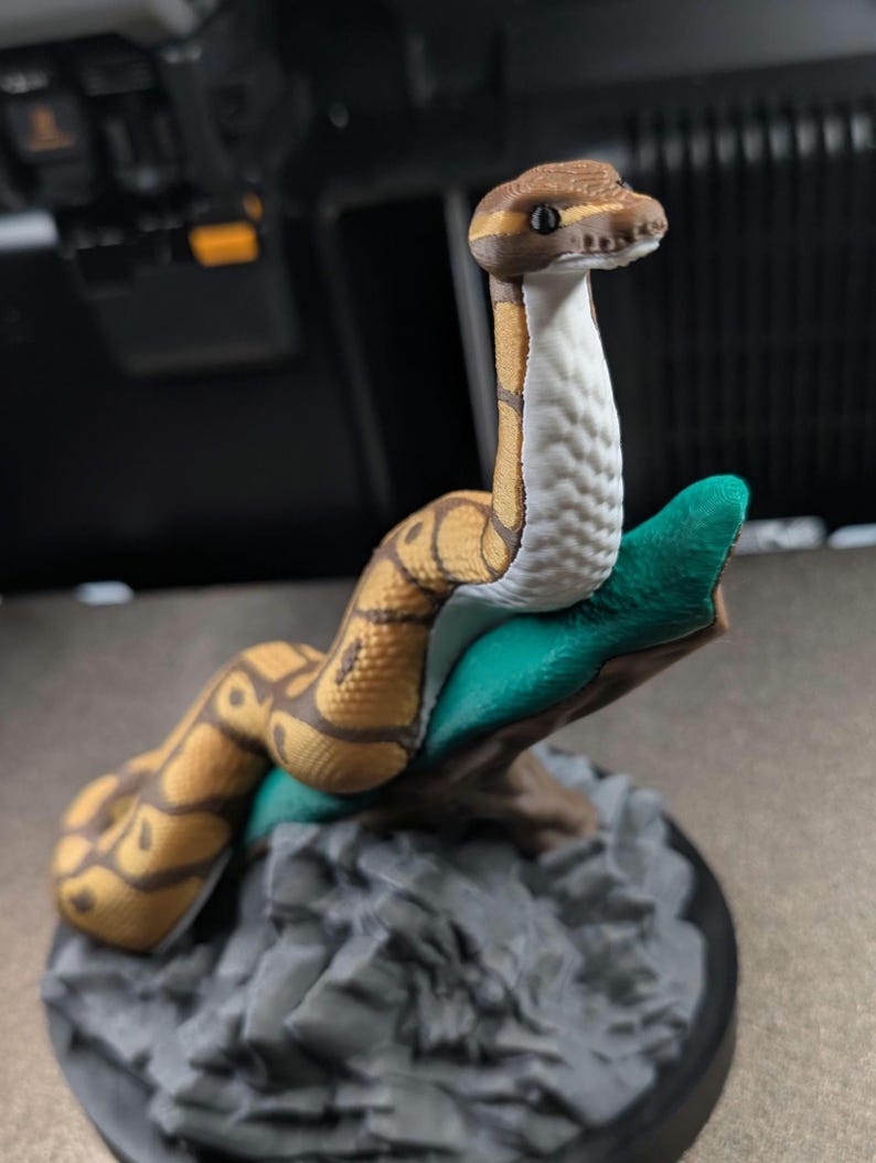 May include: A 3D printed figurine of a ball python with brown and tan patterned scales and a white underbelly. The snake is coiled around a green branch that is attached to a textured gray rock base, featuring a smooth black circular bottom.