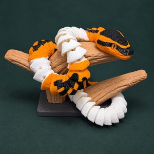 May include: A segmented, articulated snake figurine featuring a repeating pattern of white, orange, and black sections. The snake has a textured surface and is coiled around a brown, branch-like support structure mounted on a dark rectangular base.