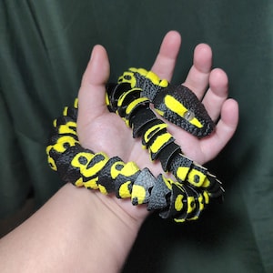 May include: A flexible, articulated toy snake featuring a textured black and bright yellow pattern. The snake has a segmented body design with a distinct head, visible eyes, and a repeating color scheme across its entire length, creating a realistic appearance.