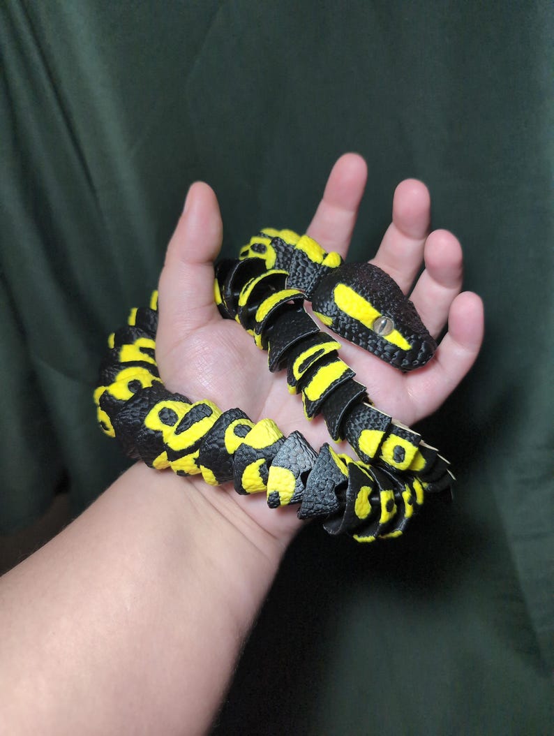 May include: A flexible, articulated toy snake featuring a textured black and bright yellow pattern. The snake has a segmented body design with a distinct head, visible eyes, and a repeating color scheme across its entire length, creating a realistic appearance.