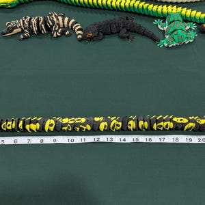 May include: A black and yellow articulated snake toy positioned above a white measuring tape that shows a length of approximately 25 inches. Several other articulated animal figures, including lizards and a frog, are arranged in the background.