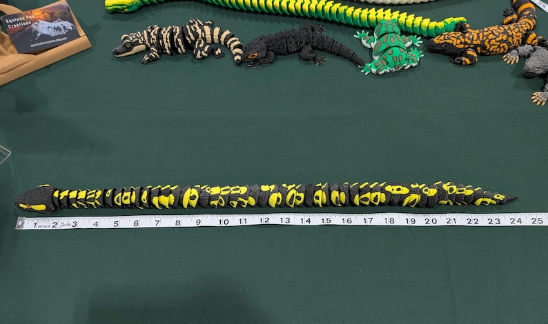 May include: A black and yellow articulated snake toy positioned above a white measuring tape that shows a length of approximately 25 inches. Several other articulated animal figures, including lizards and a frog, are arranged in the background.