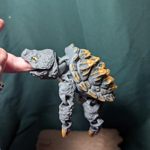 May include: A gray articulated snapping turtle figure with a textured shell featuring gold-colored accents on the ridges. The figure has a detailed head with a yellow eye, jointed limbs with pointed claws, and is positioned above a brown base.