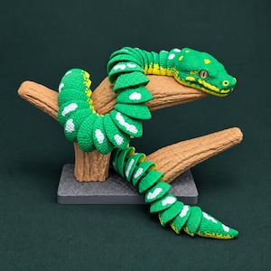 May include: A green articulated snake figurine with yellow and white markings, coiled around a textured brown branch stand. The snake features a detailed head with visible eyes and a segmented body design, mounted on a rectangular gray base.