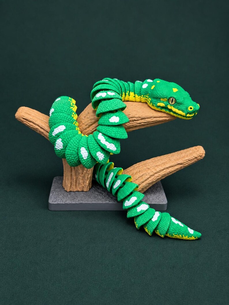 May include: A green articulated snake figurine with yellow and white markings, coiled around a textured brown branch stand. The snake features a detailed head with visible eyes and a segmented body design, mounted on a rectangular gray base.
