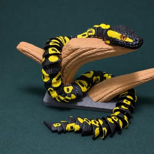May include: A segmented articulated snake figurine featuring a black and yellow patterned body with a textured surface. The snake is coiled around a light brown branch-like support structure that is mounted on a small rectangular gray base.