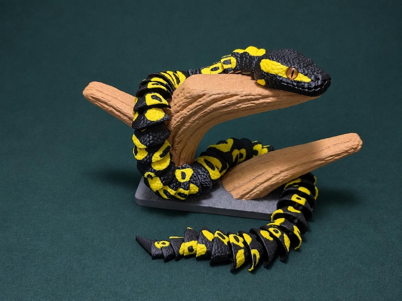 May include: A segmented articulated snake figurine featuring a black and yellow patterned body with a textured surface. The snake is coiled around a light brown branch-like support structure that is mounted on a small rectangular gray base.