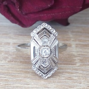 Peut inclure: Une bague en argent avec un motif géométrique orné d'un diamant central et de petits diamants tout autour. La bague est posée sur une surface en bois.
